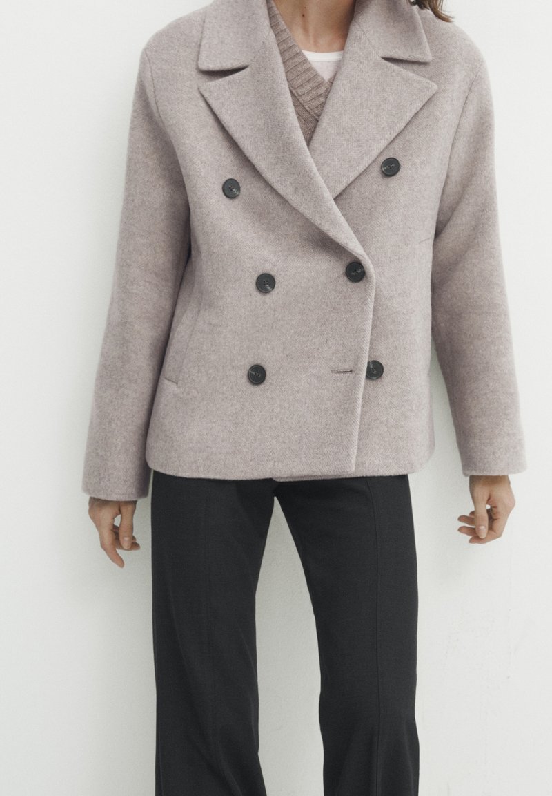 Personne portant un manteau en laine gris clair croisé avec des boutons noirs, un pull beige en dessous et un pantalon noir sur fond blanc.