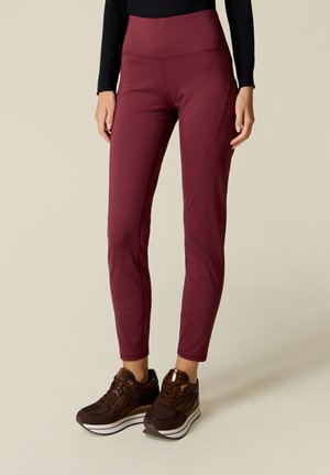 Persoon die hooggetailleerde bordeauxkleurige leggings draagt, gecombineerd met donkerbruine platformsneakers op een neutrale achtergrond.