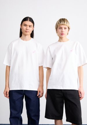 To personer iført hvide t-shirts lavet af bomuld. T-shirtsne har et pink grafik på venstre brystområde. Den ene har mørke jeans på, mens den anden har sorte shorts.