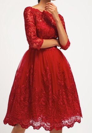 Mujer con un vestido de encaje rojo hasta la rodilla, con mangas tres cuartos, patrones florales y dobladillo festoneado, posando con una mano cerca de su rostro.