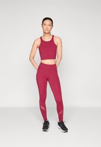 Burgundin värinen urheilullinen crop-top ja korkea vyötärö leggingsit, joissa on verkkopaneeleita, pehmeä kangas ja istuva muotoilu, yhdistettynä mustiin urheilusandaaleihin.