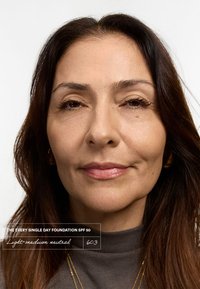 Un look de maquillage naturel présentant une peau lisse, un fond de teint neutre léger à moyen et un gloss à lèvres subtil. Le modèle a les cheveux brun foncé et porte une chemise grise.