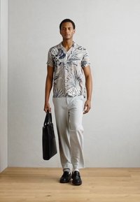 Cămașă cu mâneci scurte, pe un fundal bej deschis, cu un model floral albastru, asociată cu pantaloni gri deschis și pantofi din piele negri, purtând o geantă neagră.