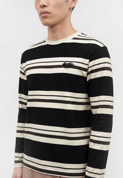 Homme portant une chemise noire et beige à manches longues avec de larges rayures horizontales et un petit logo brodé sur la poitrine, sur fond uni.