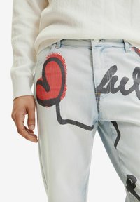 Jeans de lavado claro con un gran corazón rojo y diseños de script abstracto en negro, usados con un suéter blanco, con la mano descansando al costado.