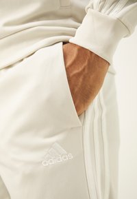 Svetlo béžové Adidas tepláky s rebrovanými manžetami, s diskrétnym logom a bočnou vreckom. Hladká tkanina s jemnou textúrou.