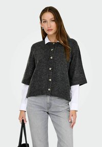 Femme portant un cardigan bouton gris foncé sur une chemise blanche et un jean gris clair, tenant un sac à main noir de la main gauche.