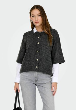 Femme portant un cardigan bouton gris foncé sur une chemise blanche et un jean gris clair, tenant un sac à main noir de la main gauche.