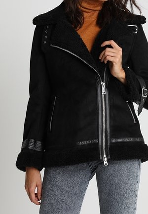 Woman wearing a black shearling jacket with silver zippers and buckle details, an orange turtleneck, and light blue jeans.