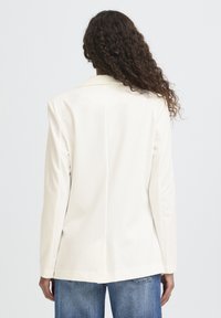 Blazer blanc en tissu lisse, doté d'un col à cran et de manches longues. Le dos présente une silhouette épurée et ajustée. Associé à un jean bleu.