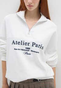 Pullover bianco con colletto con zip, caratterizzato da testo navy "Atelier Paris" e una data. Il tessuto sembra morbido e ha una vestibilità rilassata.
