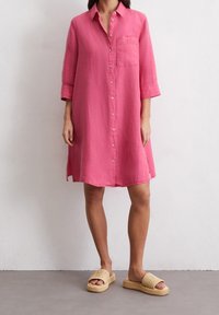 Różowa sukienka koszulowa z lnu z kołnierzykiem, zapięciem na guziki i kieszenią na piersi. Posiada rękawy o długości trzech kwartałów oraz rozcięcia po bokach dla większej swobody ruchów.