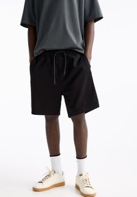Shorts en coton noir avec une ceinture élastique et un cordon de serrage, assortis à un t-shirt gris ample et des chaussettes blanches. Porte des baskets de couleur crème.