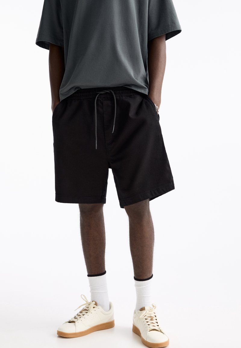 Shorts en coton noir avec une ceinture élastique et un cordon de serrage, assortis à un t-shirt gris ample et des chaussettes blanches. Porte des baskets de couleur crème.
