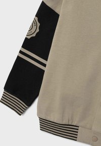 Veste pilote beige et noire avec des poignets à rayures côtelées, présentant un détail de logo et des panneaux de manches contrastants. Texture de tissu douce.