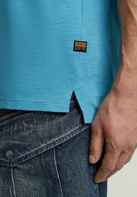 Camicia azzurra chiara a maniche corte con finitura testurizzata, caratterizzata da uno spacco laterale e un'etichetta G-Star. Indossata con jeans di denim scuro.