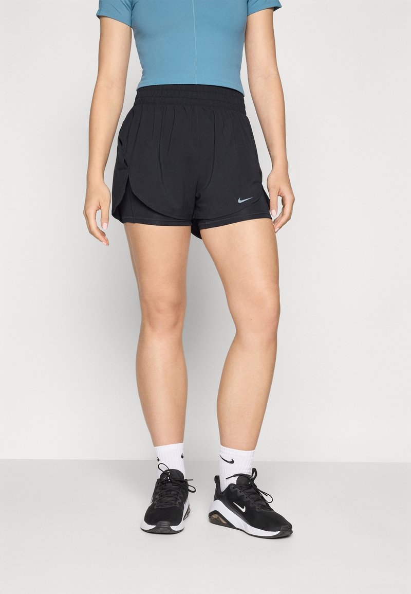 Personne portant un short Nike noir, des chaussettes Nike blanches, des baskets Nike noires et un haut bleu à manches courtes, debout sur un sol gris.