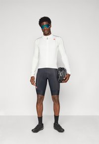 Weißes Langarm-Radsporttrikot mit Reißverschluss vorne, kombiniert mit schwarzen gepolsterten Shorts. Hält einen schwarzen Fahrradhelm. Schwarze Radschuhe.