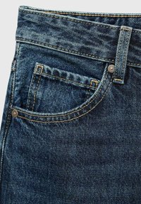 Donkerblauwe denim jeans met een zichtbare voorzak, contrasterende stiksels en een metalen knoopaccent. De textuur lijkt iets versleten.