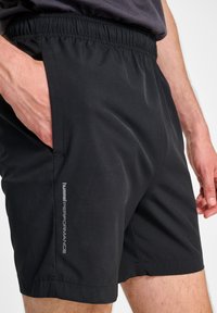 Svarta atletiska shorts med elastisk midjeband, sidofickor och en slät textur. Har en logotyp tryckt på den nedre sidan.