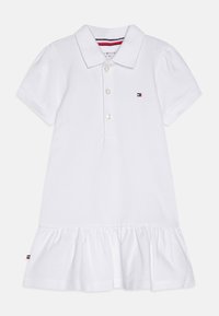 Valkoinen poolomekko, jossa on kaulus, kolme nappia ja lyhyet pörröiset hihat. Helma on viimeistelty röyhelöillä ja logotiedoilla.