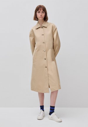 Donna con cappotto beige al ginocchio con bottoni, calzini a righe blu e neri e scarpe da ginnastica bianche, in piedi davanti a uno sfondo semplice.