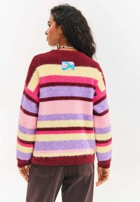 Kolorowy sweter z dzianiny w poziome paski w kolorach bordowym, różowym, lawendowym, żółtym i kremowym. Na plecach znajduje się niebieska metka.