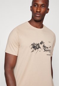 Beżowa koszulka z krótkim rękawem wykonana z miękkiego materiału, zdobiona czarnym nadrukiem górskim oraz nazwą marki "MAMMUT" na dole.