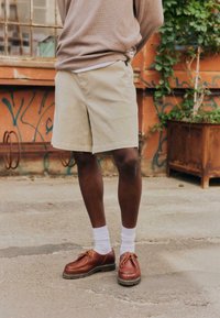 Personne portant un short beige, des chaussettes blanches et des chaussures en cuir marron à lacets, debout sur une surface en béton près d'un mur couleur rouille avec des graffitis.