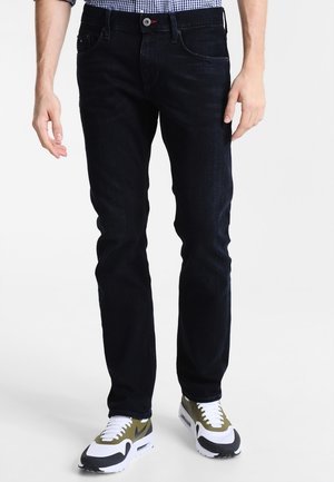 Mann trägt schwarze Slim-Fit-Jeans und weiße, schwarze sowie olivgrüne Sneakers, steht vor einem einfarbig weißen Hintergrund.