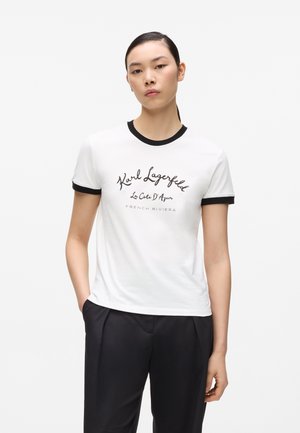Mujer joven con camiseta blanca de Karl Lagerfeld con ribete negro y pantalones negros, de pie con una mano en el bolsillo sobre fondo blanco.