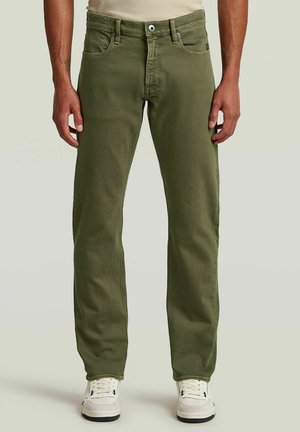 Hombre vistiendo pantalones de pierna recta color verde oliva y zapatillas blancas, de pie contra un fondo claro y liso.