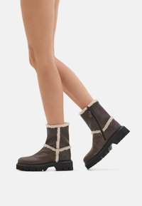 Stivaletti in pelle marrone con bordi in shearling bianco. Presentano una cerniera laterale e una robusta suola in gomma nera. Finitura testurizzata con accenti a contrasto.