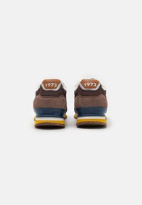 Zapatillas de ante y malla marrones con acentos en azul y amarillo, que incluyen una entresuela blanca y el año "1973" impreso en el talón.
