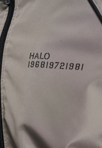 Tissu beige avec une fermeture éclair noire à gauche et le texte "HALO 196819721981" imprimé en noir sur le côté droit.