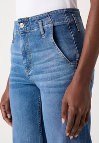 Jean en denim bleu clair avec taille haute, comportant cinq poches, des coutures visibles et une texture lisse. Fermeture à bouton à la taille.