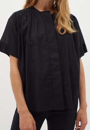 Mujer con una blusa negra de manga corta y suelta, con un escote fruncido y una tapeta frontal oculta, combinada con pantalones negros.