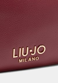 Bordová texturovaná kožená taška s embosovaným logem "LIU·JO MILANO" v metalické zlaté barvě. Hladký povrch s jemným vzorováním.