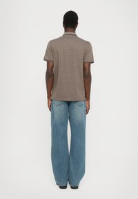 Brun mönstrad polo med korta ärmar, kontrasterad av mörka detaljer, tillsammans med ljusblå vida jeans. Svarta skor synliga.