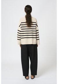 Beżowy sweter z dzianiny w czarne paski, luźny krój, długie rękawy, zestawiony z czarnymi plisowanymi spodniami i czarnymi płaskimi sandałami, widziany od tyłu.