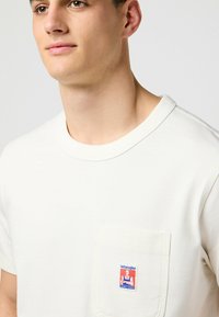 Hvid crew neck t-shirt med korte ærmer, der har en lomme på venstre bryst og et blåt og rødt "Wrangler"-mærke på lommen.