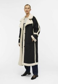 Schwarzer langer Mantel mit cremefarbener Kunstfellfütterung, großem Kragen, Knopfverschluss und Taschen. Verfügt über kontrastierende Streifen entlang der Ärmel.