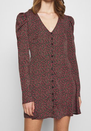 Femme portant une robe boutonnée à manches longues avec un petit motif floral noir, rouge et vert et un décolleté en V.