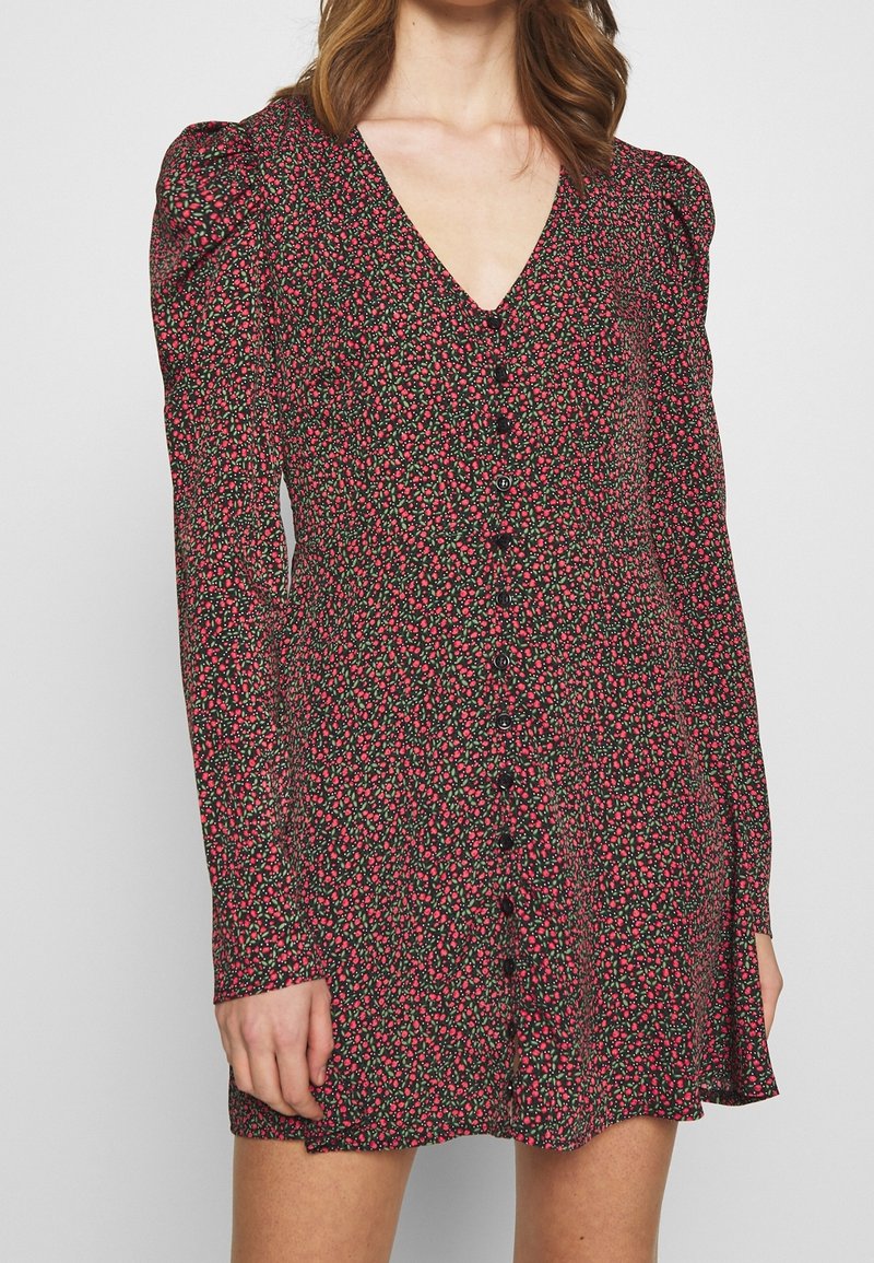 Femme portant une robe boutonnée à manches longues avec un petit motif floral noir, rouge et vert et un décolleté en V.
