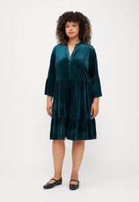 Tīklu zamšādas kleita ar V veida kakla izgriezumu un trīs ceturtdaļu zvana piedurknēm, ar vairākiem slāņiem un mīkstu struktūru. Valkāta ar melnām plakanām kurpēm.