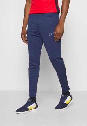 Persoon draagt marineblauwe Nike trainingsbroek met een lichtblauw logo, zwarte sokken, marineblauwe en gele Nike-sneakers en een rood shirt.