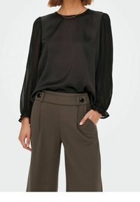 Kvinna klädd i en svart genomskinlig blus med långa ärmar som är instoppad i high-waist stora bruna byxor med knapppdetaljer på midjan.