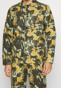 Grön-och-gul mönstrad jacka i canvas med knappstängning framtill, krage och två framfickor. Har en logotyp-patch på vänster bröst.