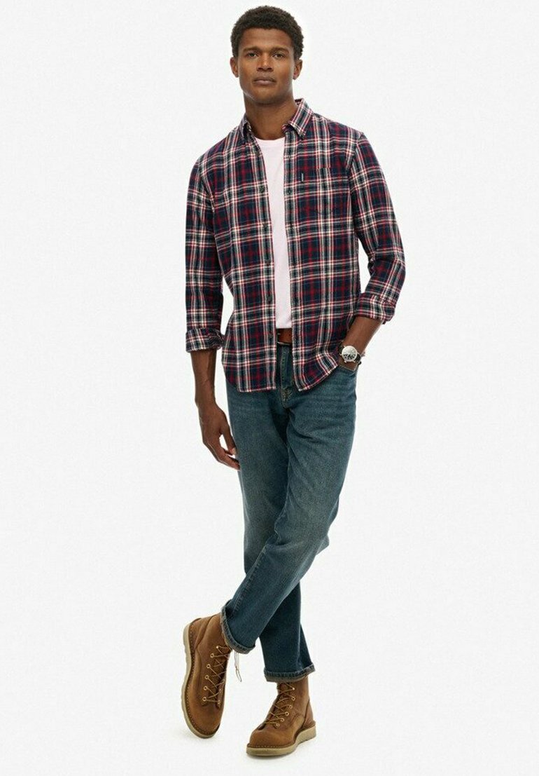 Camisa de franela a cuadros en azul marino y rojo, llevada sobre una camiseta clara, combinada con jeans oscuros y botas marrones con cordones. Look casual.