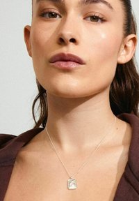 Collar de plata con un colgante rectangular que presenta un diseño texturizado. La modelo lleva una blusa marrón oscuro, mostrando un tono de piel suave.